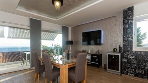 Un coin repas avec une table et des chaises en verre, un téléviseur à écran plat fixé au mur et une vue sur la mer grâce à de grandes fenêtres.