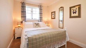 A bedroom with a bed, two bedside lamps, a window with curtains, and two framed butterfly pictures on the wall. - Armstrong Cottages No22 (Bamburgh)
