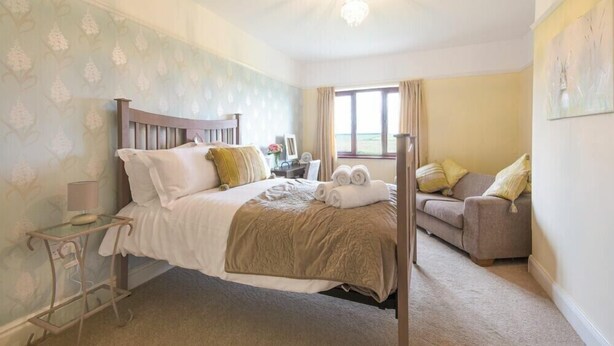 A bedroom with a four-poster bed, a bedside table with a lamp, a sofa, and a large window with curtains. - Dairyman's Cottage (Morpeth)
