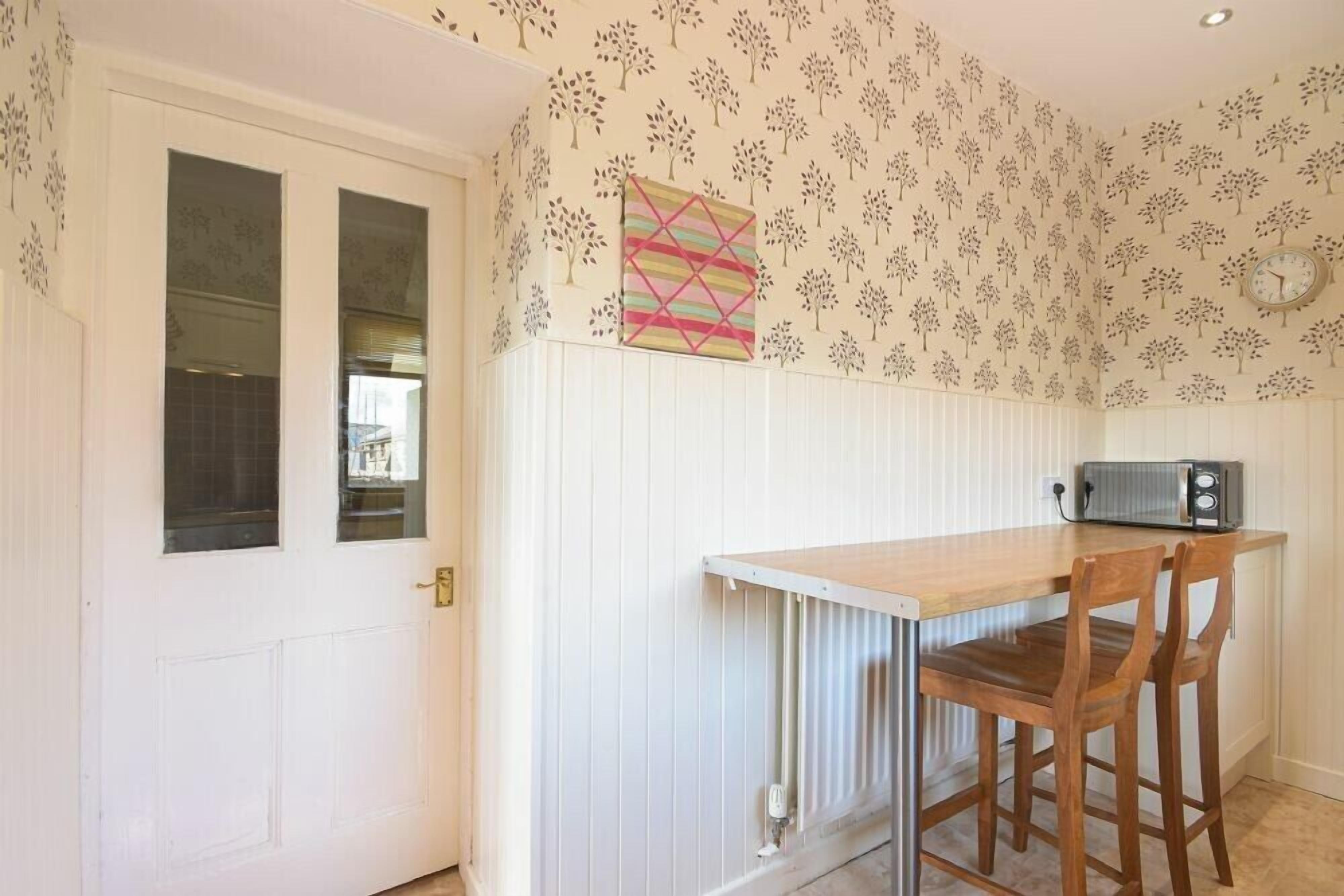 Een kleine keuken met een witte deur, een raam, een radiator, een houten tafel en twee stoelen.