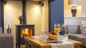 Een gezellige woonkamer met een houtkachel, een bank, een bijzettafeltje met ontbijtartikelen en een lamp.