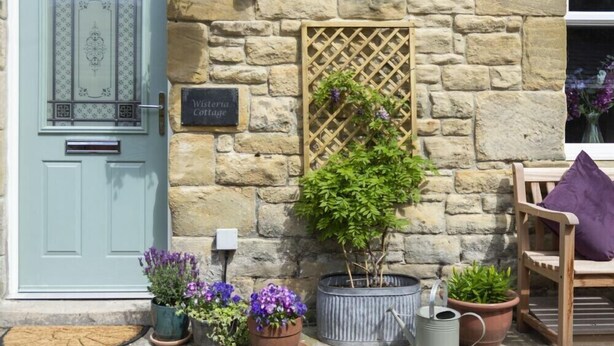 Een stenen gebouw met een lichtblauwe deur, een bord met de tekst 'Westminster Cottage' en diverse bloempotten.