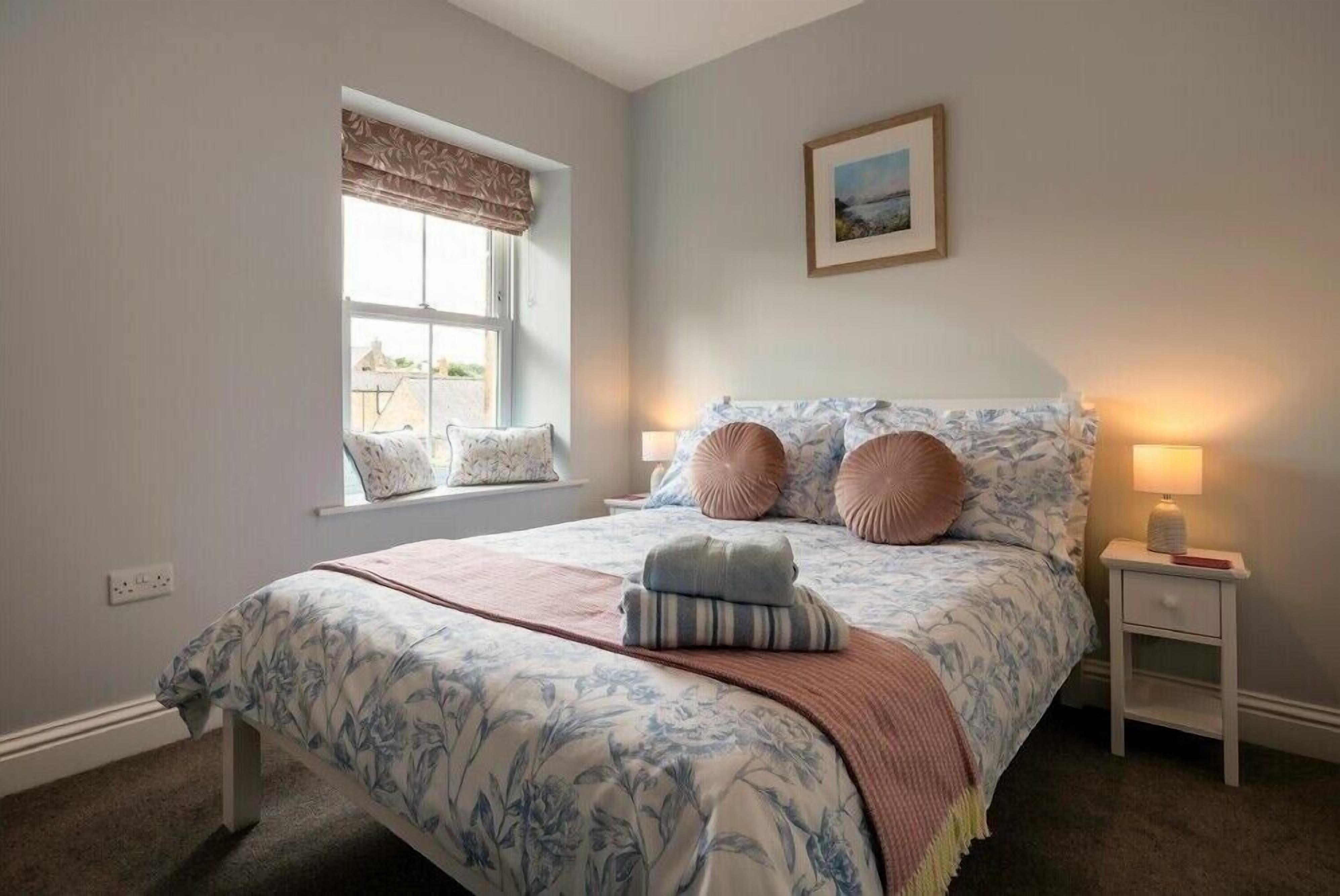 A bedroom with a large bed, a window with curtains, a bedside table with a lamp, and a framed picture on the wall.