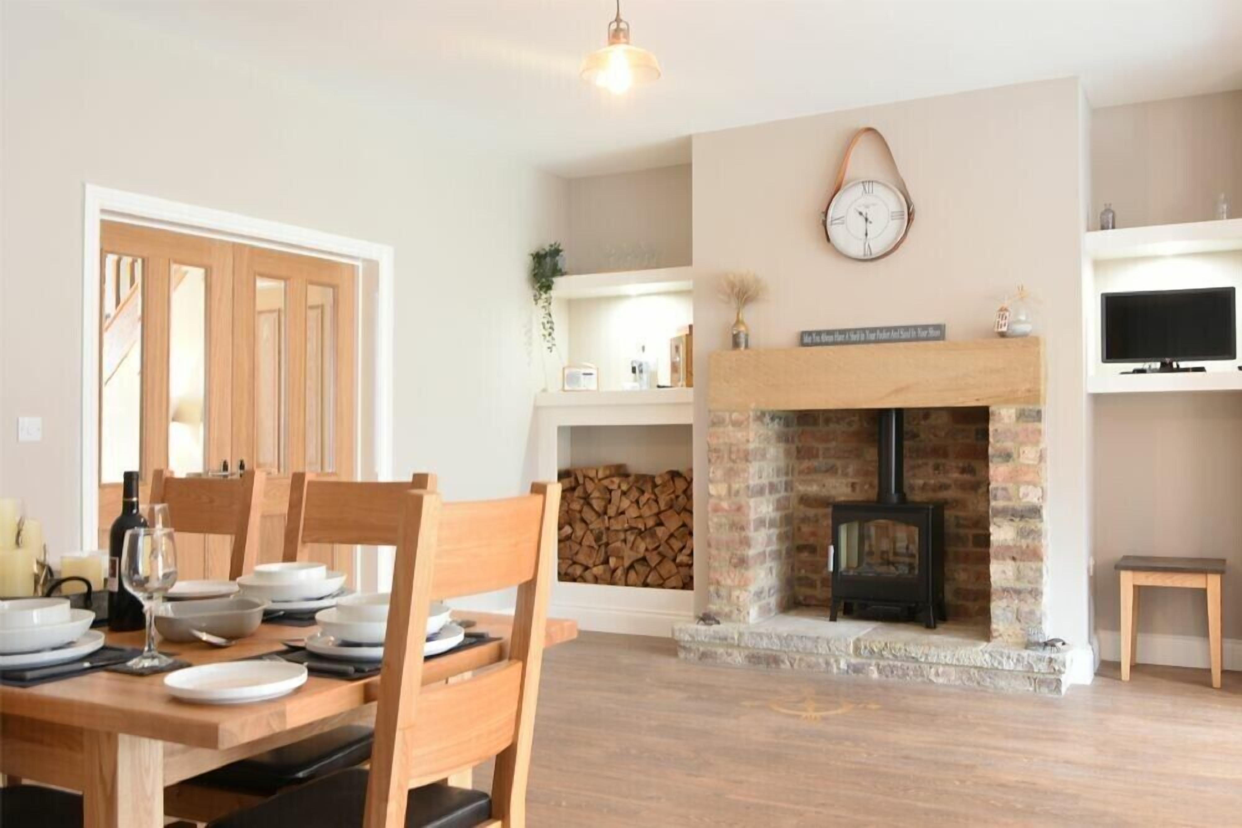 A dining area with a wooden table set for four, a fireplace with a brick surround, and a clock on the wall.