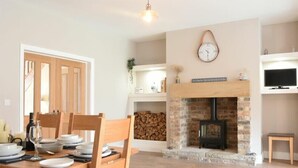 Un coin repas avec une table en bois dressée pour quatre personnes, une cheminée avec un manteau en briques et une horloge au mur.