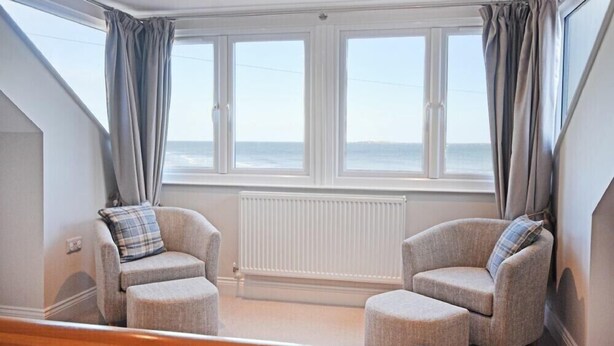 Une pièce comprenant deux fauteuils et deux poufs, un radiateur blanc et de grandes fenêtres avec des rideaux.