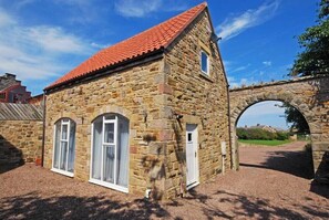A stone building with a red-tiled roof, arched windows, and a stone archway. - Quarry Mews (Bamburgh)