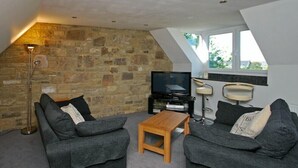 A cozy living room with two grey sofas, a wooden coffee table, a stone wall, a TV, and a window with a view.