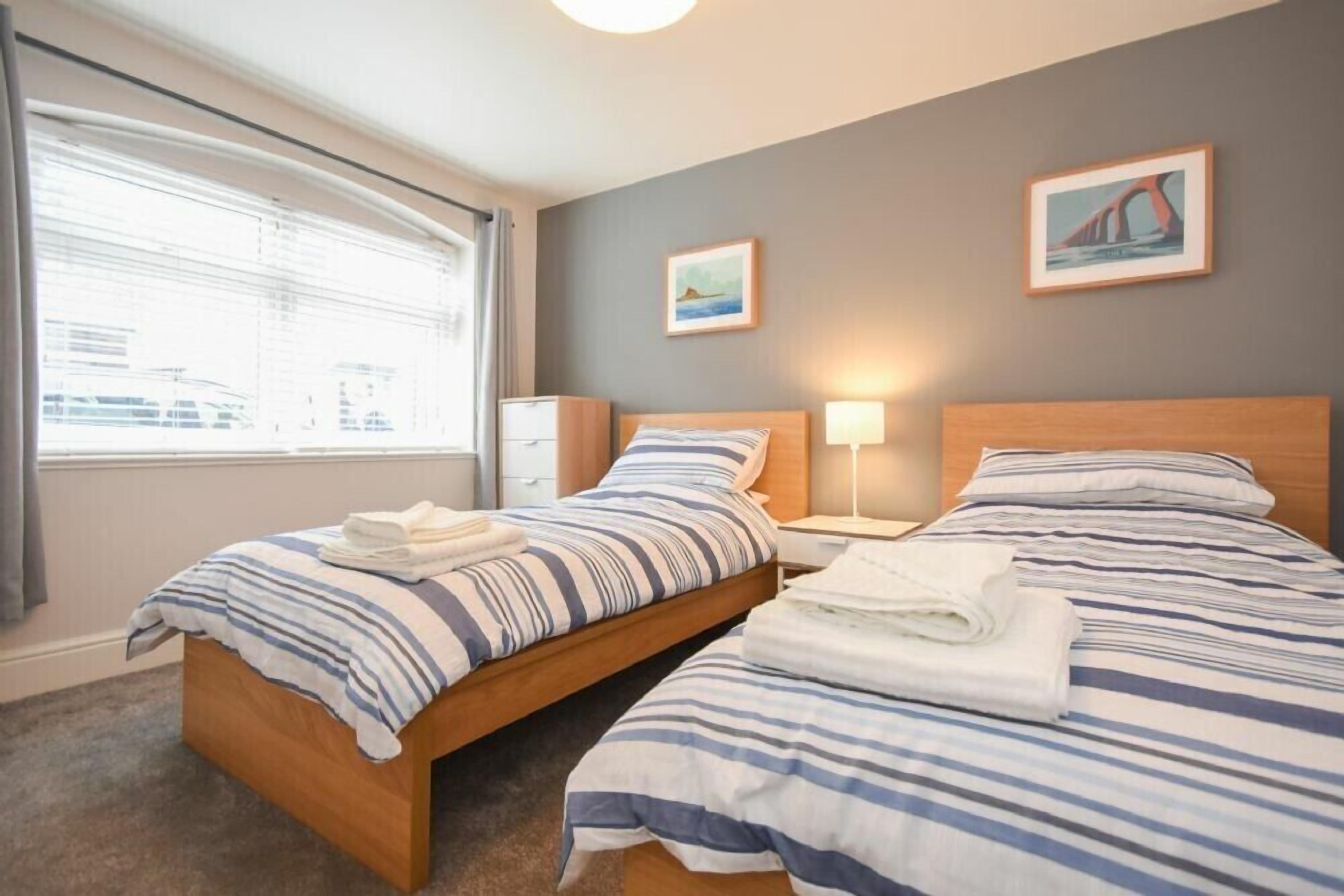 Une chambre d’hôtel avec deux lits, une fenêtre avec des stores, une table de chevet avec une lampe et deux tableaux encadrés au mur.