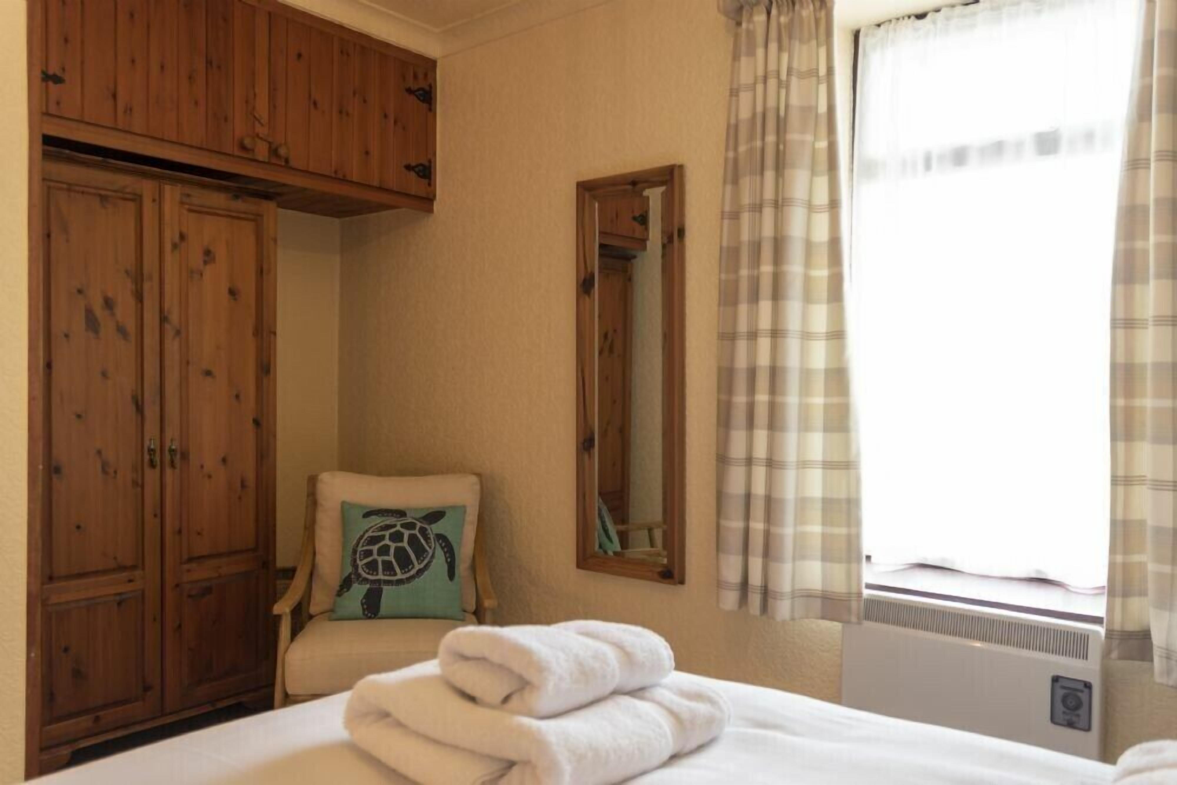 A bedroom with a bed, a chair with a pillow, a wooden wardrobe, a mirror, and a window with curtains.