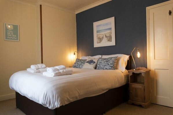 A neatly made bed with white linens and a few decorative pillows, a bedside table with a lamp, and a framed picture on the wall. - Kittiwake Cottage, Seahouses (Seahouses)