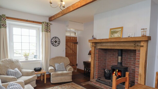 A cozy living room with a fireplace, a sofa set, a wooden table, and a large window with curtains. - No2 Budle Bay Cottage (Bamburgh)