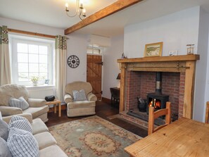 A cozy living room with a fireplace, a sofa set, a wooden table, and a large window with curtains. - No2 Budle Bay Cottage (Bamburgh)