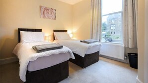 A hotel room with two beds, a window with curtains, a radiator, and a wall-mounted light fixture. - Sunnybrae (Alnwick)