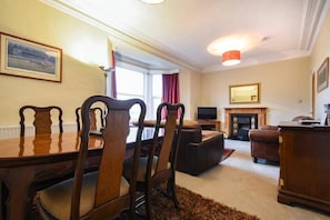 A dining area with a wooden table and chairs, a fireplace, and a television in a living room with a sofa and a fireplace mantel.