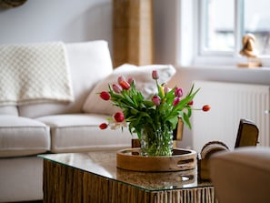 A living room with a glass coffee table, a vase with flowers, a wooden side table, and a white sofa.