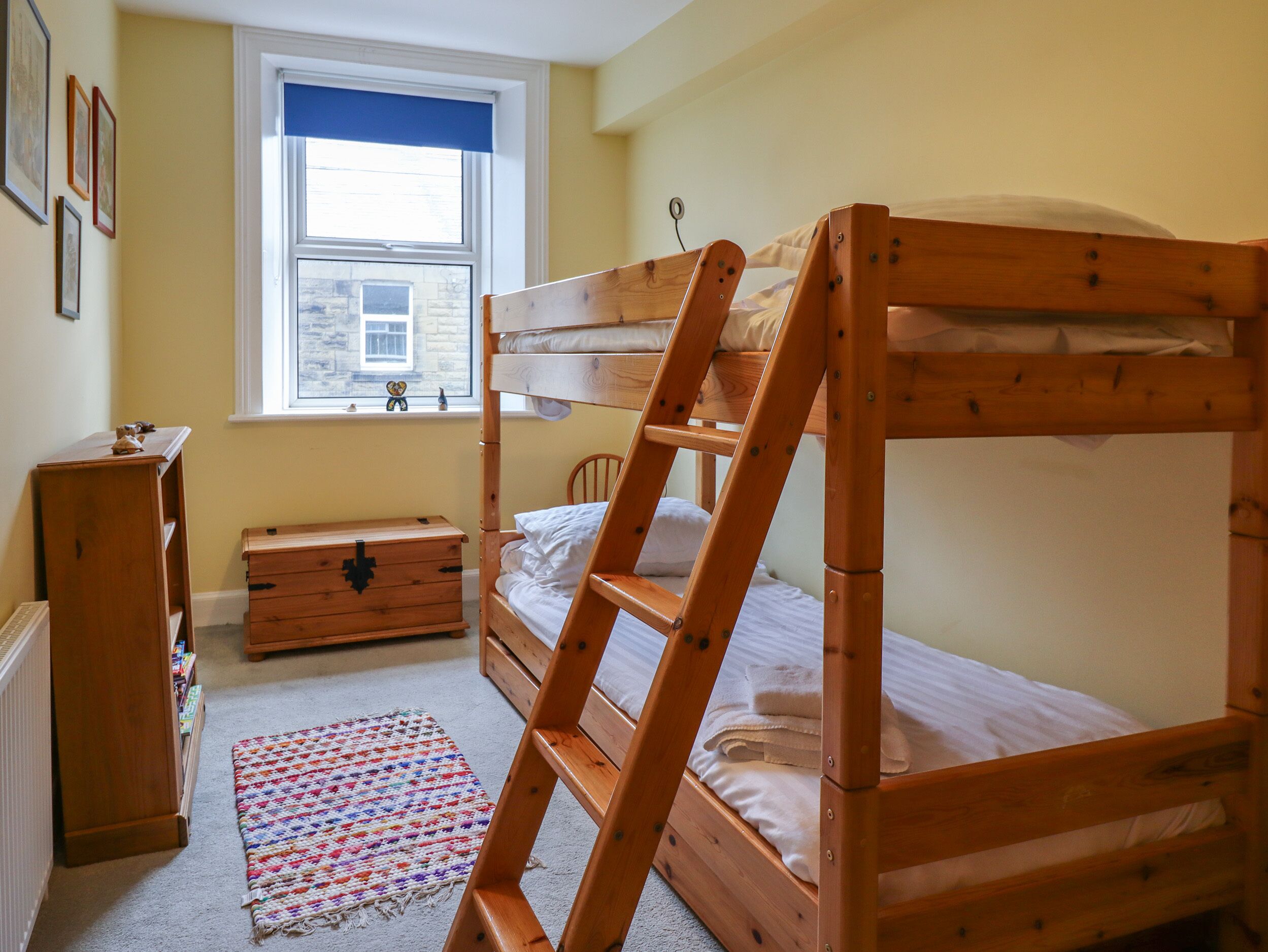 A bunk bed with a ladder, a wooden dresser, a small chest of drawers, a colorful rug, and a window with a view of a stone building.