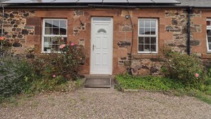 A stone building with a white door, two windows, and a solar panel roof. - Budle Bay Cottage (Bamburgh)