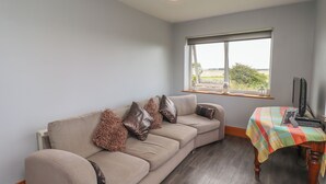 A room with a beige sofa, a wooden table with a plaid tablecloth, and a window with a view of the outdoors. - Budle Bay Cottage (Bamburgh)