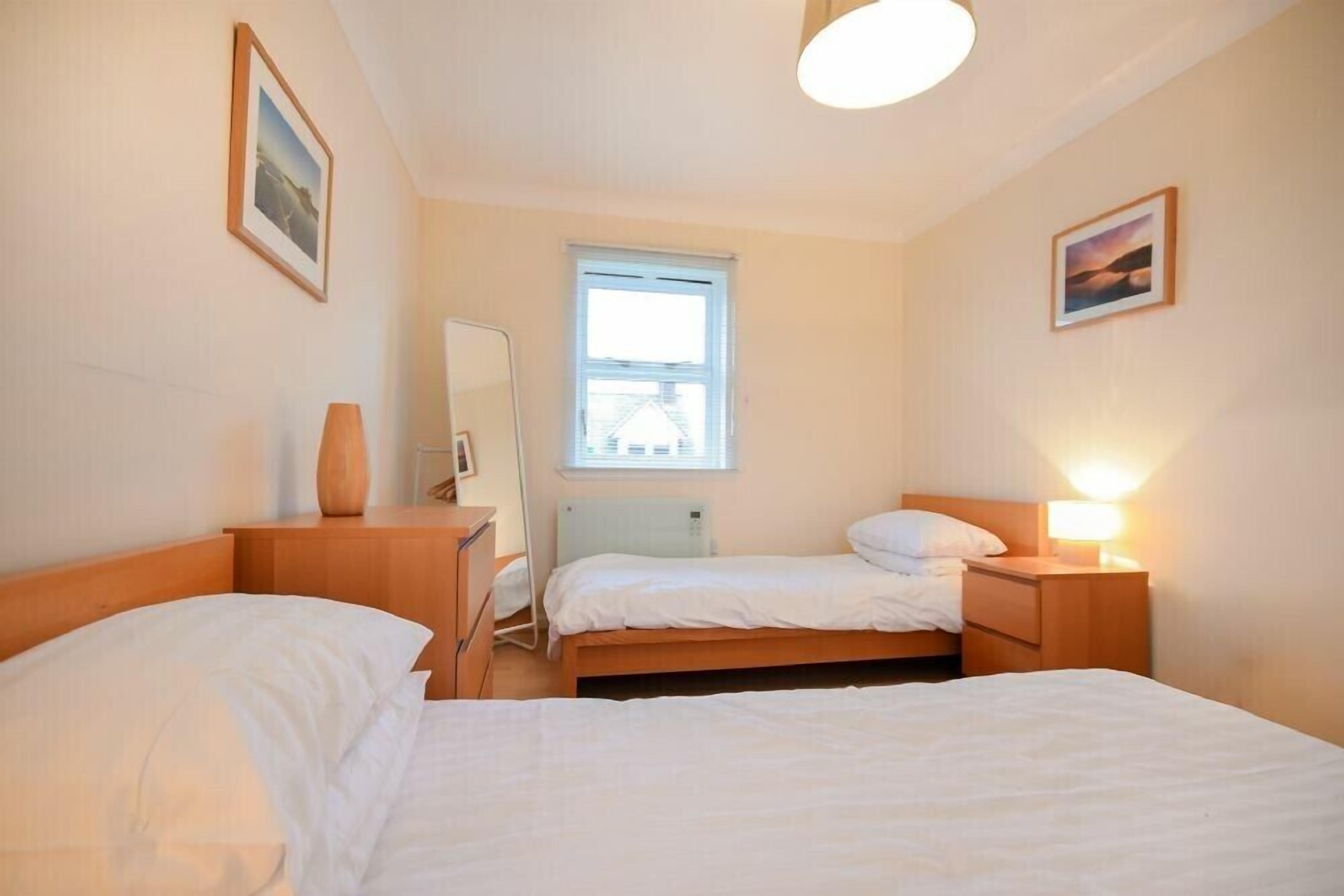 Ein Schlafzimmer mit zwei Betten, einem Nachttisch mit Lampe, einer Kommode, einem Fenster mit Jalousien und einem gerahmten Bild an der Wand.