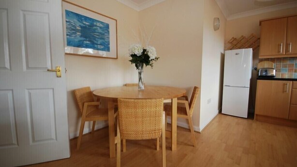 Un coin repas avec une table ronde et quatre chaises, une porte blanche, un tableau encadré, un vase avec des fleurs et une cuisine dotée de meubles en bois et d’un réfrigérateur.