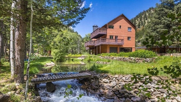 Um edifício de dois andares com varanda, cercado por uma paisagem natural com um riacho e árvores.