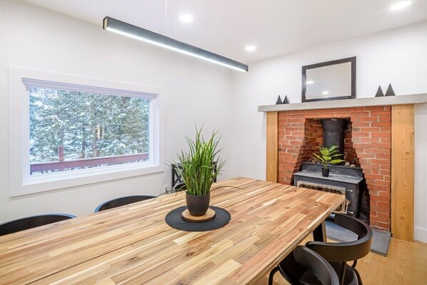 Un espace repas moderne avec une table en bois, des chaises noires et une plante en pot. On aperçoit une cheminée en briques avec une cheminée en arrière-plan.