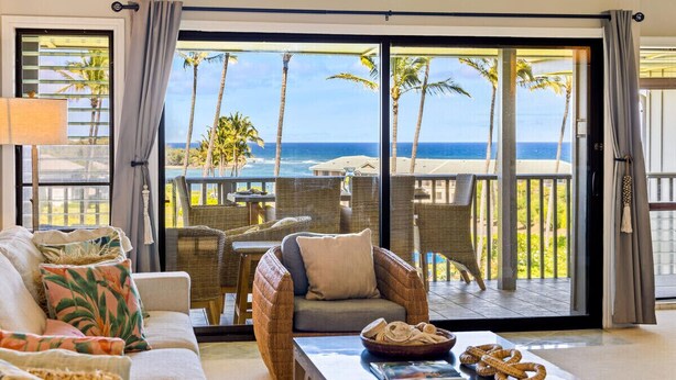 A room with a sofa, wicker chairs, a table with a bowl of bread, and a view of the ocean through large glass doors. - KAUAI KONG QUARTERS OCEANVIEW 2 Bdrm 2 KINGS 1 TWIN. NextDoor2Hyatt Dining&Bars. (Koloa)