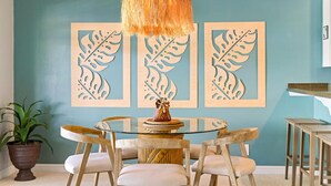 A dining area with a glass table surrounded by wooden chairs, a hanging light fixture, and framed leaf artwork on the wall. - KAUAI KONG QUARTERS OCEANVIEW 2 Bdrm 2 KINGS 1 TWIN. NextDoor2Hyatt Dining&Bars. (Koloa)
