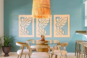 A dining area with a glass table surrounded by wooden chairs, a hanging light fixture, and framed leaf artwork on the wall.