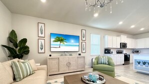 A modern living room with a sofa, a coffee table, a TV displaying a beach scene, and a kitchen in the background.