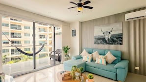 Una sala de estar moderna con un sofá azul, una mesa de centro y una hamaca junto a un ventanal con vistas a un edificio.
