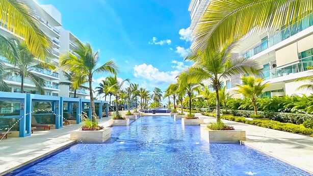Un hotel moderno con una piscina central, rodeado de palmeras y balcones de vidrio.