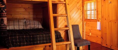Interior de una cabaña de madera con literas, una escalera, una silla y una ventana.