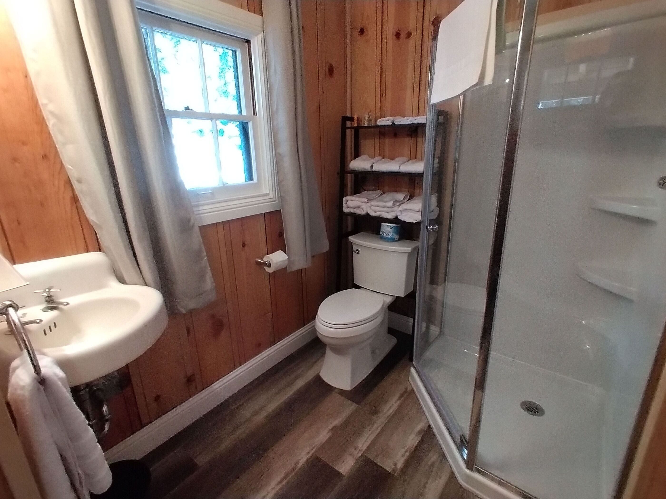 A small bathroom with a wooden floor, a white sink, a toilet, and a glass-enclosed shower.