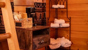 A wooden cabin room with a wooden bed, a small wooden table with a basket, a coffee maker, and a shelf with towels. - Lakeshore Resort (Lakeshore)