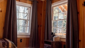 A room with wooden paneled walls, two windows with curtains, a bed, and a small wooden nightstand with a lamp and a basket. - Lakeshore Resort (Lakeshore)