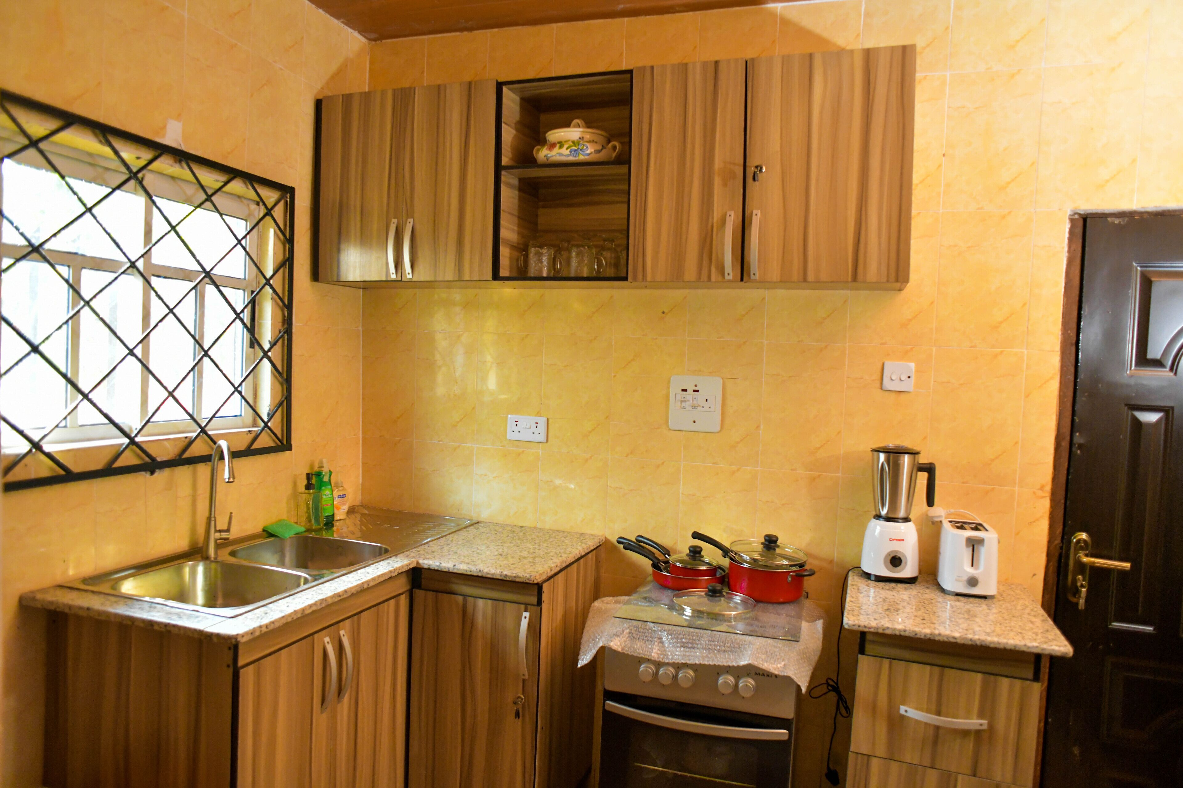 Une cuisine de taille réduite, dotée de meubles en bois, d’une cuisinière, d’un évier et d’une fenêtre avec une grille en métal.
