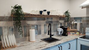 A kitchen with a marble countertop, a sink, a coffee maker, and a shelf with glassware and plants. - Cozy basement suite near the mountains (Cochrane)
