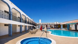 Zona de piscina amb gandules i ombrelles, cel blau clar i un edifici amb parets blaves i blanques.