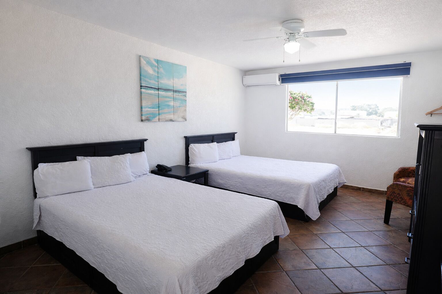 Habitación de hotel con dos camas, ventilador de techo, ventana con vistas y un cuadro en la pared.