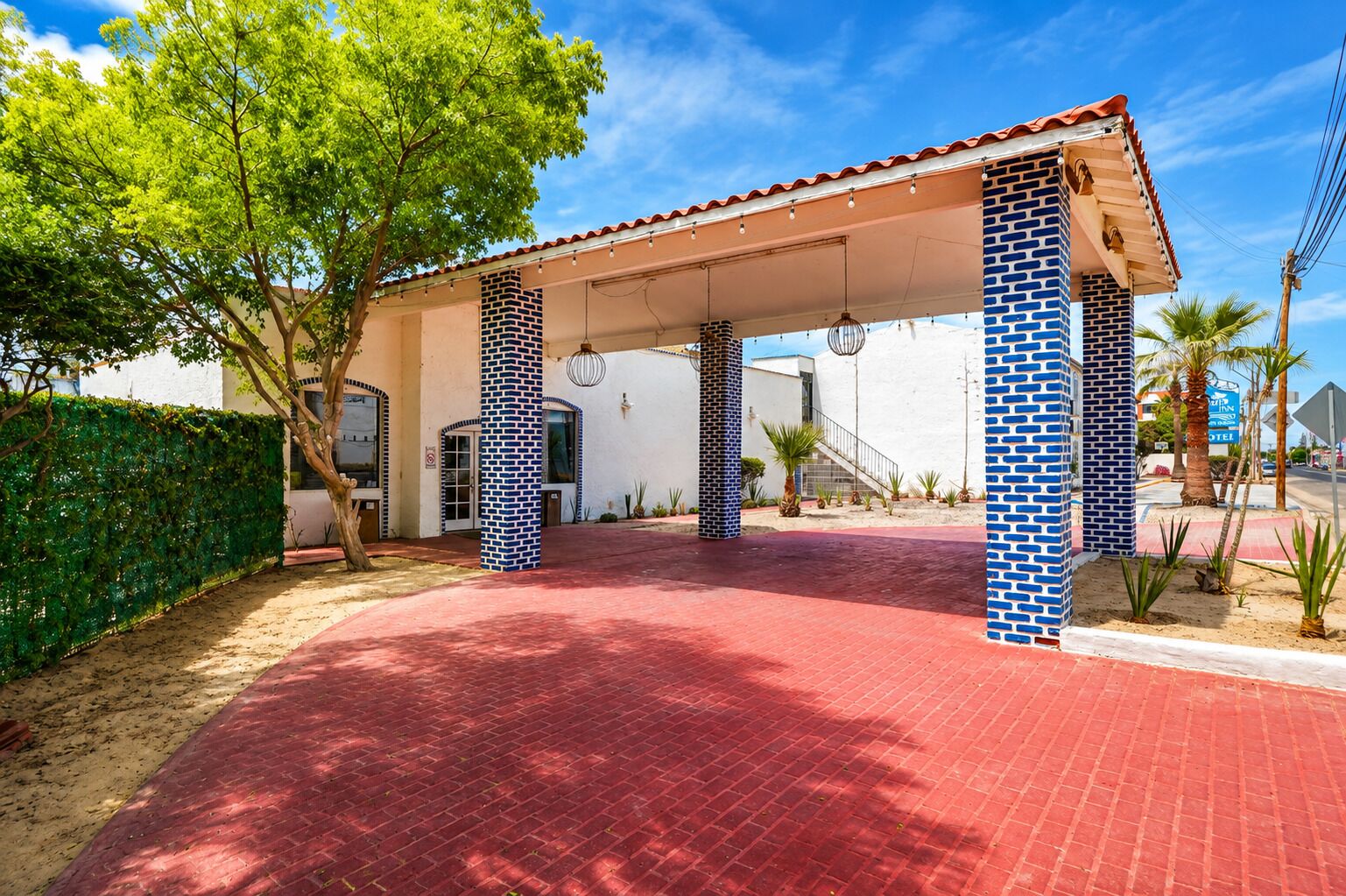 Un espacio al aire libre cubierto con piso de ladrillo rojo, columnas con diseños azules y blancos, y una palmera.