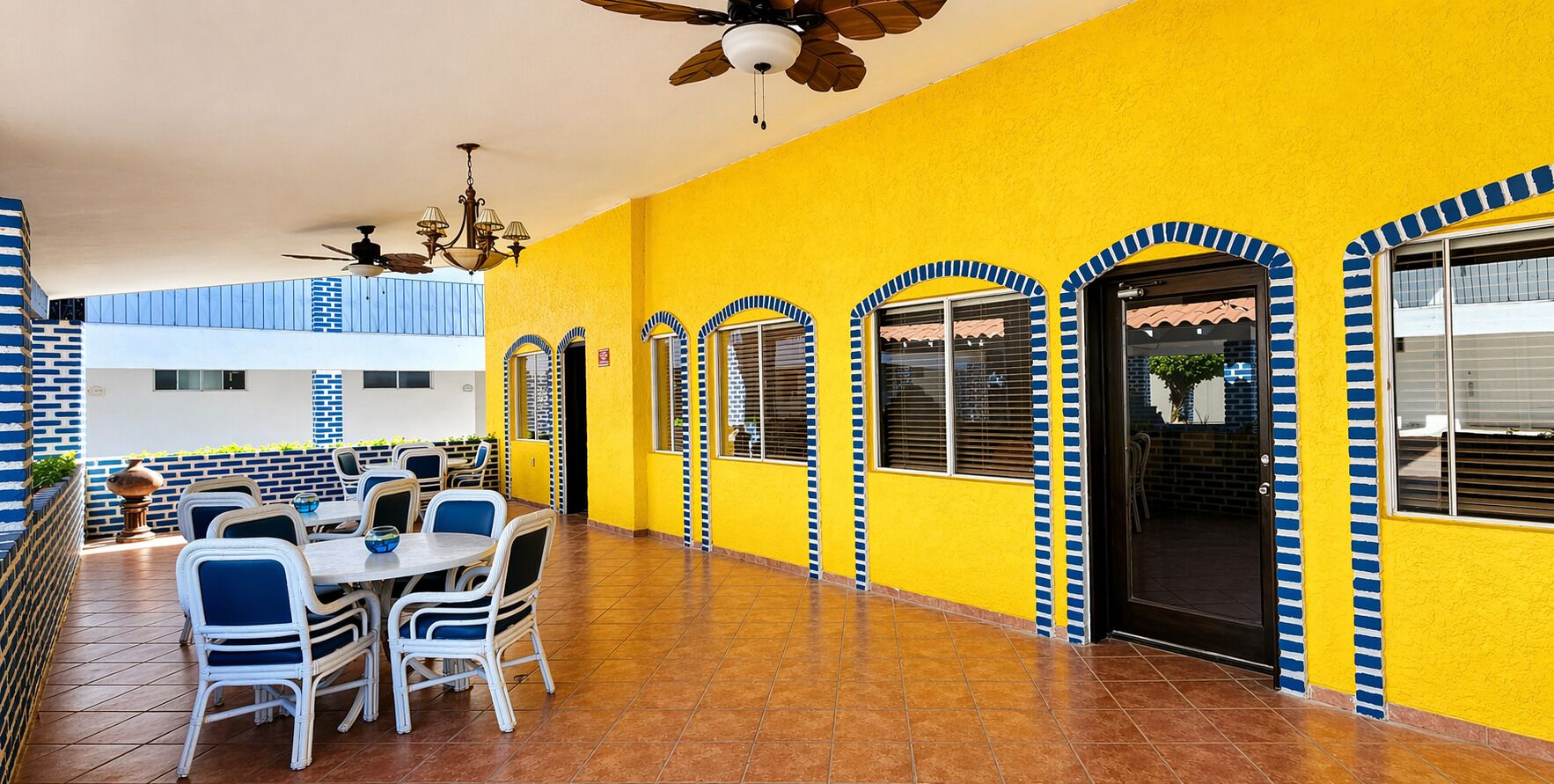 A yellow building with blue and white accents, a covered patio with tables and chairs, and a ceiling fan.