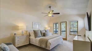 A bedroom with a bed, bedside tables, a desk, a ceiling fan, and windows with a view of the outdoors.