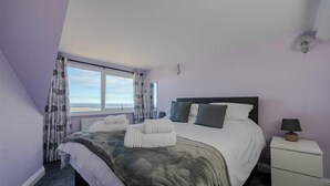 Quarto com cama, mesa de cabeceira, abajur e vista para o mar pela janela.