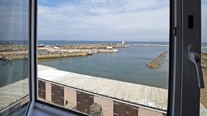 Vista da janela sobre um porto com barcos, um farol e edifícios industriais.