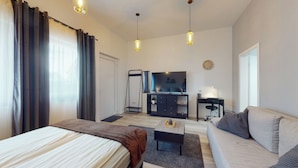 Ein modernes Hotelzimmer mit Bett, Sofa, Schreibtisch und Fernseher. Es gibt ein Fenster mit Vorhängen, eine Wanduhr und eine Pendelleuchte.