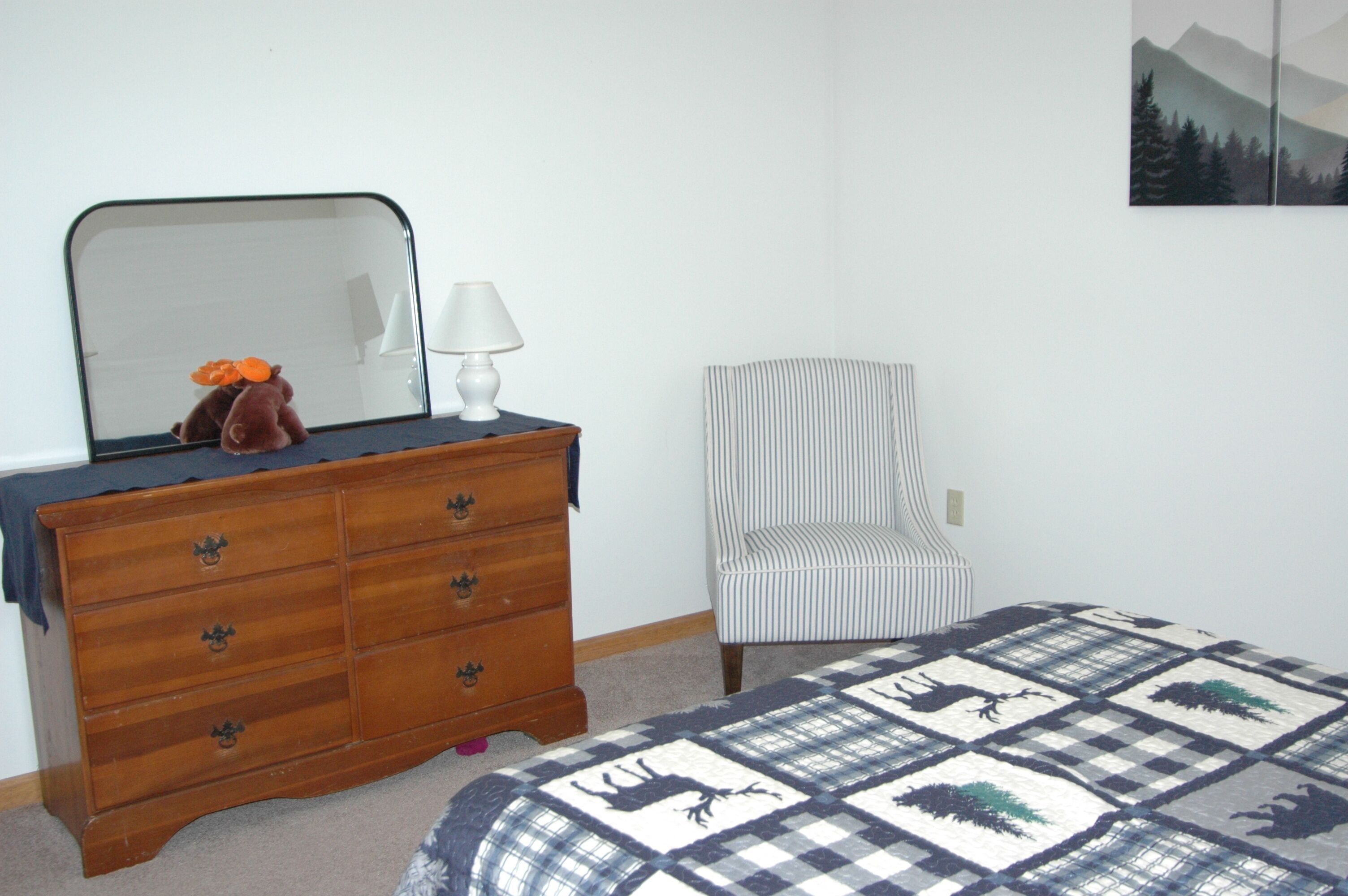 A bedroom with a wooden dresser, a chair, a bed with a checkered quilt, a lamp, and a wall-mounted mirror.