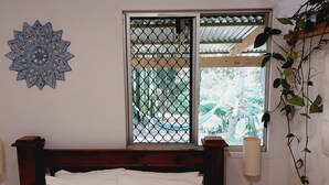 Un dormitorio con cama de madera, almohadas blancas, un adorno en la pared, una ventana con mosquitero y una lámpara de noche.