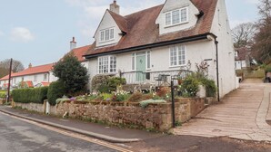 A traditional two-story house with a red-tiled roof, a stone wall with a flower bed, and a paved driveway. - Rosedale Lodge (Alnwick)